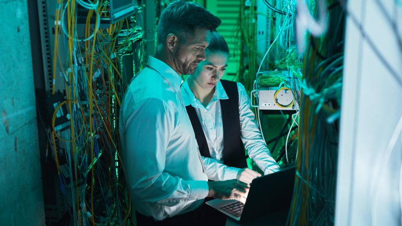 High angle view at two network technicians using laptop in server room while working with supercomputer in data center